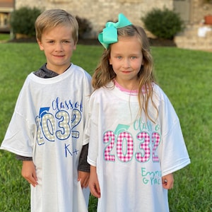 May include: Two children wearing white t-shirts with the text "Class of 2032" in blue and gray checkered print. The girl's shirt has pink and white checkered print and the boy's shirt has gray and white checkered print. The girl's shirt also has the text "Emma Grace" in blue.