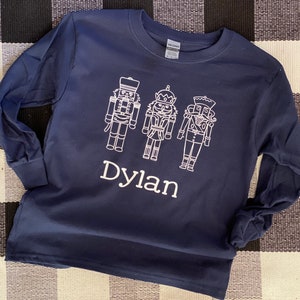 May include: A navy blue long-sleeved t-shirt with a white design of three nutcracker figures and the name "Dylan" underneath.