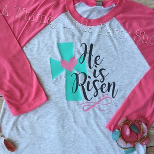May include: A grey and pink baseball tee shirt with a teal cross and a pink heart. The shirt has the text "He is Risen" in black cursive lettering.