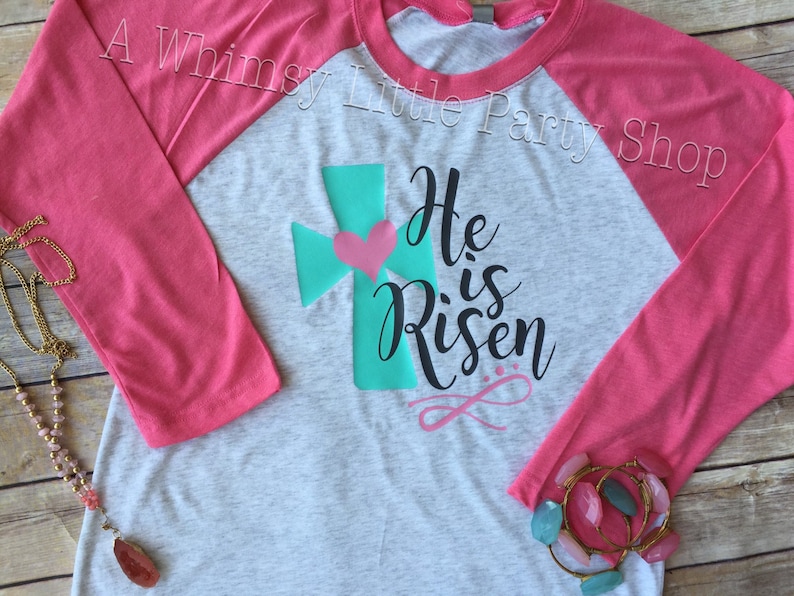 May include: A grey and pink baseball tee shirt with a teal cross and a pink heart. The shirt has the text "He is Risen" in black cursive lettering.
