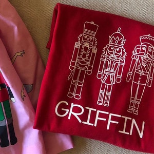 May include: Red t-shirt with a white design of three nutcracker figures and the name "GRIFFIN" printed below.