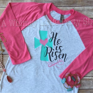 May include: A grey and pink baseball tee shirt with a teal cross and a pink heart. The shirt has the text "He is Risen" printed on it.