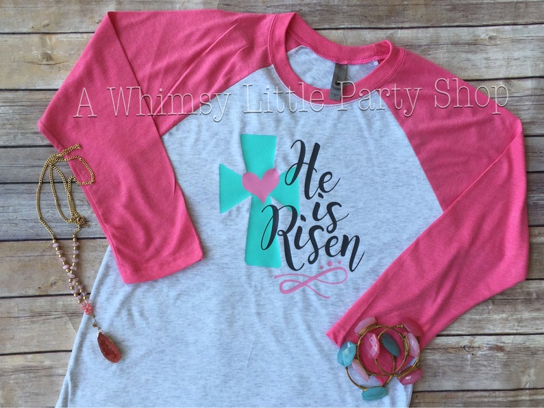 May include: A grey and pink baseball tee shirt with a teal cross and a pink heart. The shirt has the text "He is Risen" printed on it.