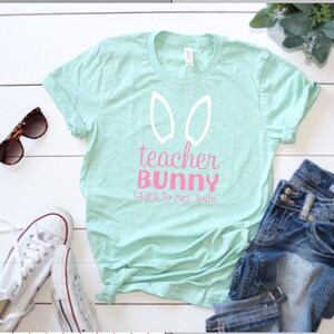 May include: A light blue t-shirt with a white bunny ear design and the text "teacher bunny" in pink. The text "well, mrs. smith" is written in black below the bunny ears.