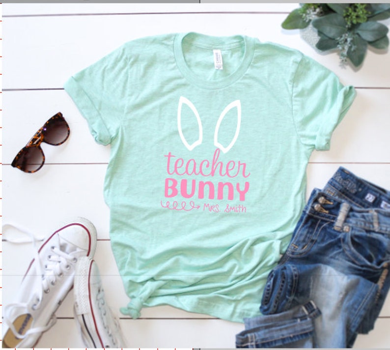 May include: A light blue t-shirt with a white bunny ear design and the text "teacher bunny" in pink. The text "well, mrs. smith" is written in black below the bunny ears.