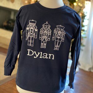 May include: A navy blue long-sleeved t-shirt with a white outline of three nutcracker figures and the name "Dylan" printed on the front.