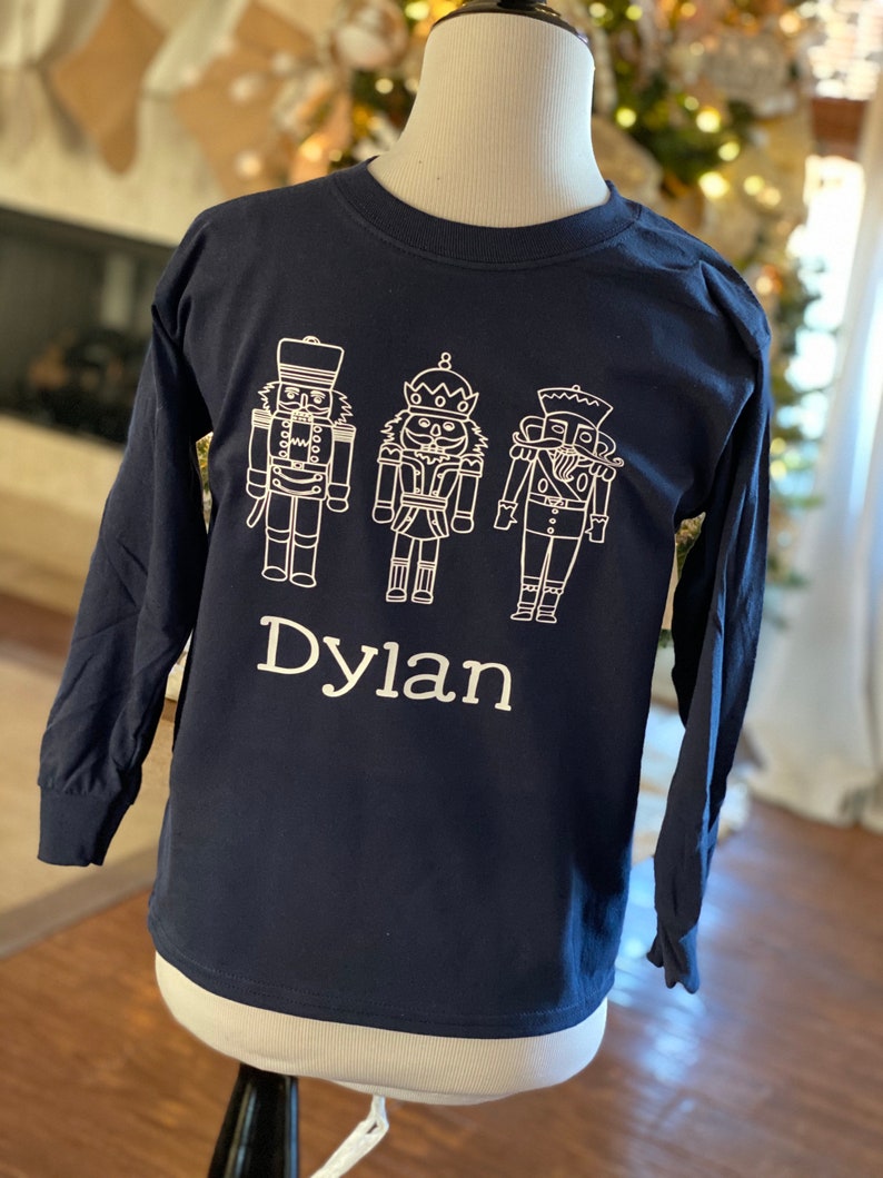 May include: A navy blue long-sleeved t-shirt with a white outline of three nutcracker figures and the name "Dylan" printed on the front.