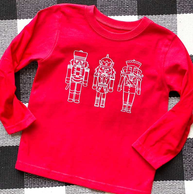 May include: Red long-sleeved shirt with a white outline of three nutcracker figures.