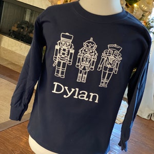 May include: A navy blue long-sleeved shirt with a white design of three nutcracker figures and the name "Dylan" printed below.