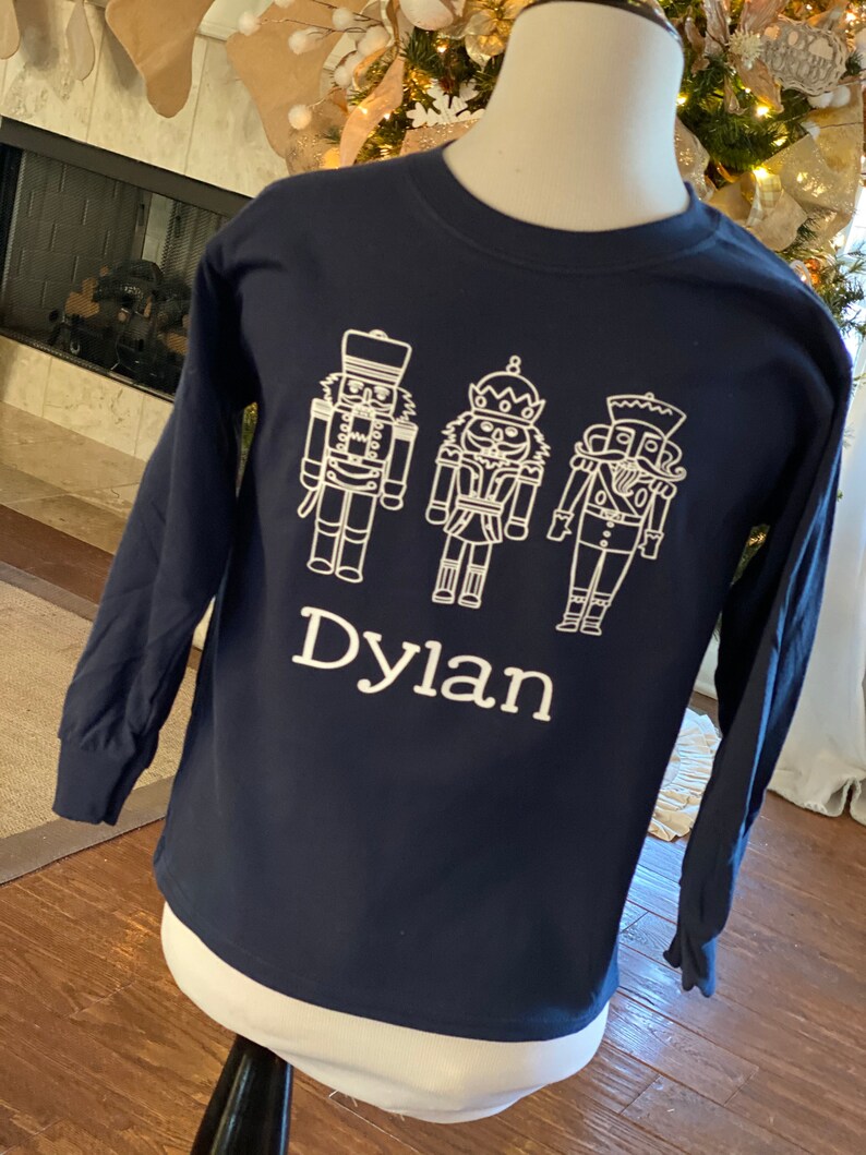 May include: A navy blue long-sleeved shirt with a white design of three nutcracker figures and the name "Dylan" printed below.