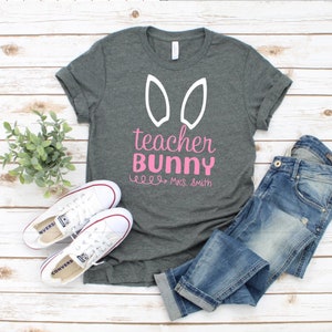 May include: A gray t-shirt with white bunny ears and the text "teacher bunny" in pink lettering. The text "Mrs. Smith" is also included in pink lettering.
