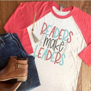 May include: A heather gray and coral raglan shirt with the text "Readers Make Leaders" in a colorful design. The image also includes blue jeans, brown leather ankle boots, and a gold necklace with a tassel.
