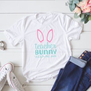 May include: White t-shirt with pink bunny ears and the text "teacher bunny" in teal blue. The text "Mrs. Smith" is written in smaller teal blue letters below the main text.