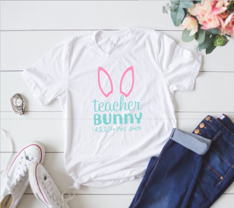 May include: White t-shirt with pink bunny ears and the text "teacher bunny" in teal blue. The text "Mrs. Smith" is written in smaller teal blue letters below the main text.