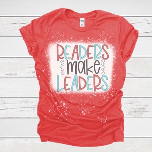 May include: A red t-shirt with the words "READERS make LEADERS" in a colorful design. The text is in a white rectangle with a distressed, speckled effect. The shirt has rolled-up sleeves.