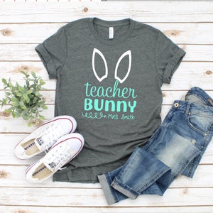 May include: A dark gray t-shirt with white bunny ears and the text "teacher bunny" in teal. The text "Mrs. Smith" is written in black below the main text.
