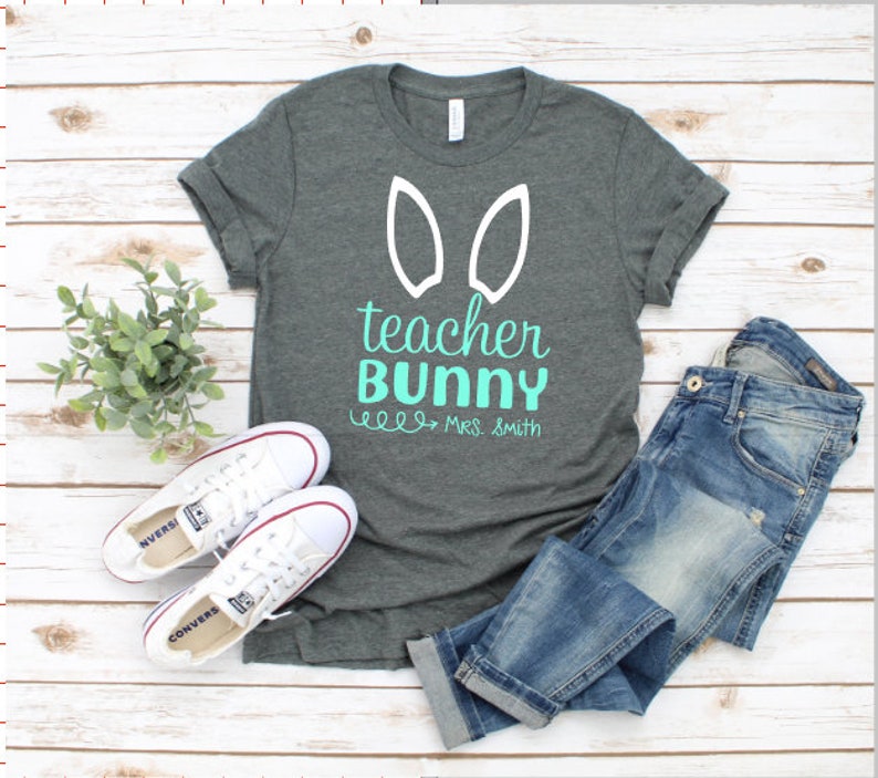 May include: A dark gray t-shirt with white bunny ears and the text "teacher bunny" in teal. The text "Mrs. Smith" is written in black below the main text.