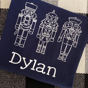 May include: A navy blue t-shirt with a white design of three nutcracker figures. The name "Dylan" is written in white below the figures.