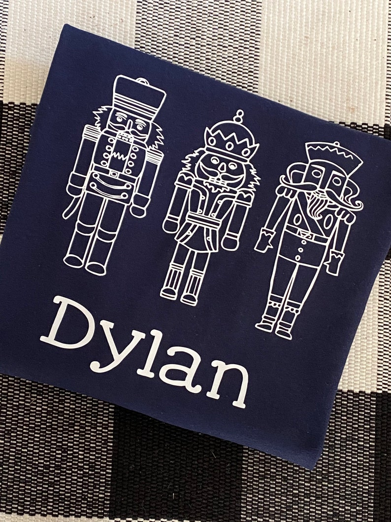 May include: A navy blue t-shirt with a white design of three nutcracker figures. The name "Dylan" is written in white below the figures.