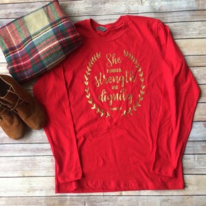 Puede incluir: Camiseta roja de manga larga con un dise&ntilde;o en l&aacute;mina dorada que dice "Ella est&aacute; vestida de fuerza y dignidad" con una corona dorada alrededor del texto.