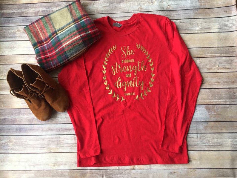 Puede incluir: Camiseta roja de manga larga con un dise&ntilde;o en l&aacute;mina dorada que dice "Ella est&aacute; vestida de fuerza y dignidad" con una corona dorada alrededor del texto.