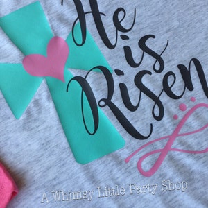 May include: A grey t-shirt with a teal cross and a pink heart. The text "He is Risen" is written in black cursive lettering.