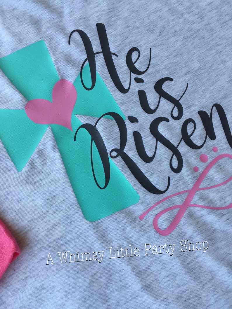 May include: A grey t-shirt with a teal cross and a pink heart. The text "He is Risen" is written in black cursive lettering.