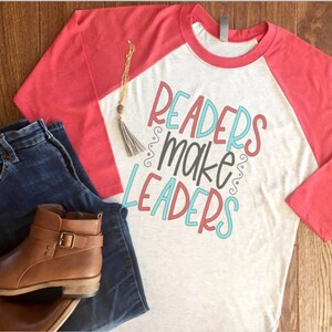 May include: A heather gray and coral raglan shirt with the text "Readers Make Leaders" in a colorful design. The image also includes blue jeans, brown leather ankle boots, and a gold necklace with a tassel.