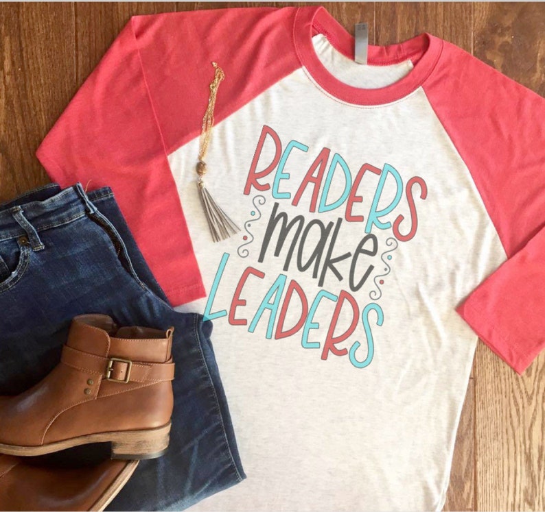 May include: A heather gray and coral raglan shirt with the text "Readers Make Leaders" in a colorful design. The image also includes blue jeans, brown leather ankle boots, and a gold necklace with a tassel.