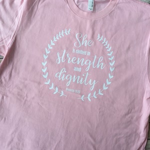 Puede incluir: Camiseta rosa con un dise&ntilde;o gr&aacute;fico blanco que dice "Ella est&aacute; vestida de fuerza y dignidad" con una corona alrededor del texto y las palabras "Proverbios 31:25" debajo de la corona.