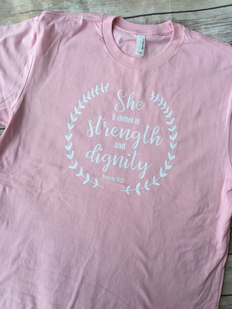 Puede incluir: Camiseta rosa con un dise&ntilde;o gr&aacute;fico blanco que dice "Ella est&aacute; vestida de fuerza y dignidad" con una corona alrededor del texto y las palabras "Proverbios 31:25" debajo de la corona.