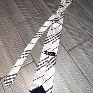 May include: A white tie with a black and gray plaid pattern. The tie has a label that says "Burberry". The tie is made of a silky material.