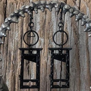 May include: Two black guillotine earrings hanging from a decorative metal arch. Each earring features a guillotine design with a blade and frame. The earrings are suspended from a chain and a circular ring. The arch is textured and has a dark, metallic finish, set against a wooden background.
