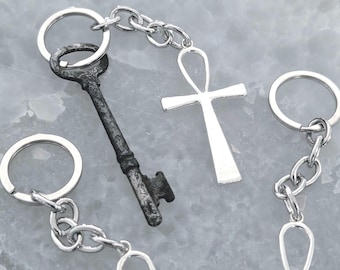 Pode incluir: Uma coleção de porta-chaves de metal prateado com símbolos ankh e uma chave antiga escura com aspeto desgastado. Cada peça está presa a um anel circular por uma corrente curta de prata, mostrando o contraste entre o acabamento polido dos ankhs e a aparência texturizada e envelhecida da chave.