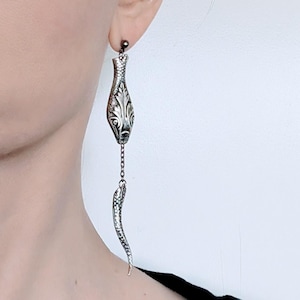 May include: A silver-toned earring featuring a detailed snake design. The earring has a long, dangling element with a snake's body and head, connected by a chain to a decorative top piece. The design includes intricate patterns and textures.