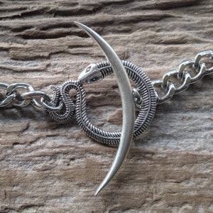 May include: A silver chain bracelet with a snake and crescent moon charm. The snake is coiled around the crescent moon, and the charm is attached to the chain with a silver ring.