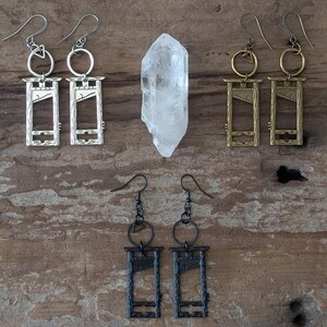 May include: An assortment of handmade earrings with a guillotine design in silver, gold, and black. A clear quartz crystal is also present. The earrings are displayed on a rustic wooden surface.