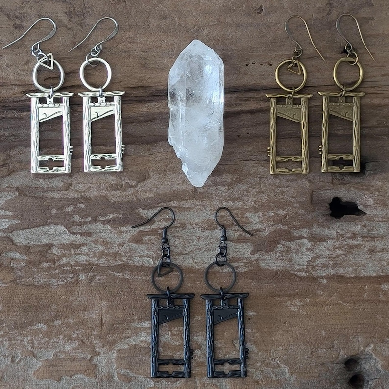 May include: An assortment of handmade earrings with a guillotine design in silver, gold, and black. A clear quartz crystal is also present. The earrings are displayed on a rustic wooden surface.
