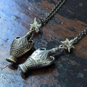 Könnte beinhalten: Zwei silberne und bronzene Halsketten mit komplizierten Designs. Jede Halskette verfügt über einen kleinen, detaillierten, verzierten Flaschenanhänger und einen Knochenanhänger.