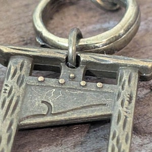 May include: A detailed close-up of a bronze-colored pendant featuring a miniature guillotine design. The pendant has a textured surface and is attached to a circular bail. The guillotine has small, round rivets and a curved blade.