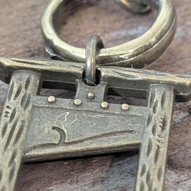 May include: A detailed close-up of a bronze-colored pendant featuring a miniature guillotine design. The pendant has a textured surface and is attached to a circular bail. The guillotine has small, round rivets and a curved blade.