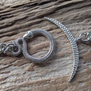 May include: A silver snake charm with a textured body and a crescent moon charm with a textured surface. Both charms are attached to a silver chain.