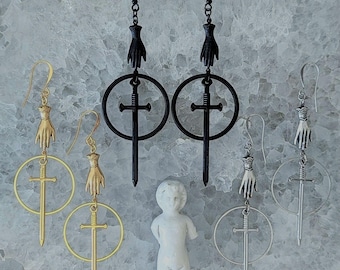 May include: Three pairs of dangle earrings with a sword and hand design. The earrings are in gold, silver, and black.