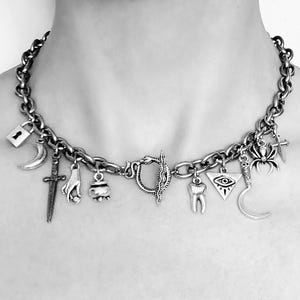 Puede incluir: Un collar de cadena de plata con múltiples dijes, incluyendo una luna creciente, un candado, una daga, una mano, un caldero, una serpiente, un diente, un triángulo con un ojo, una araña y una cruz.