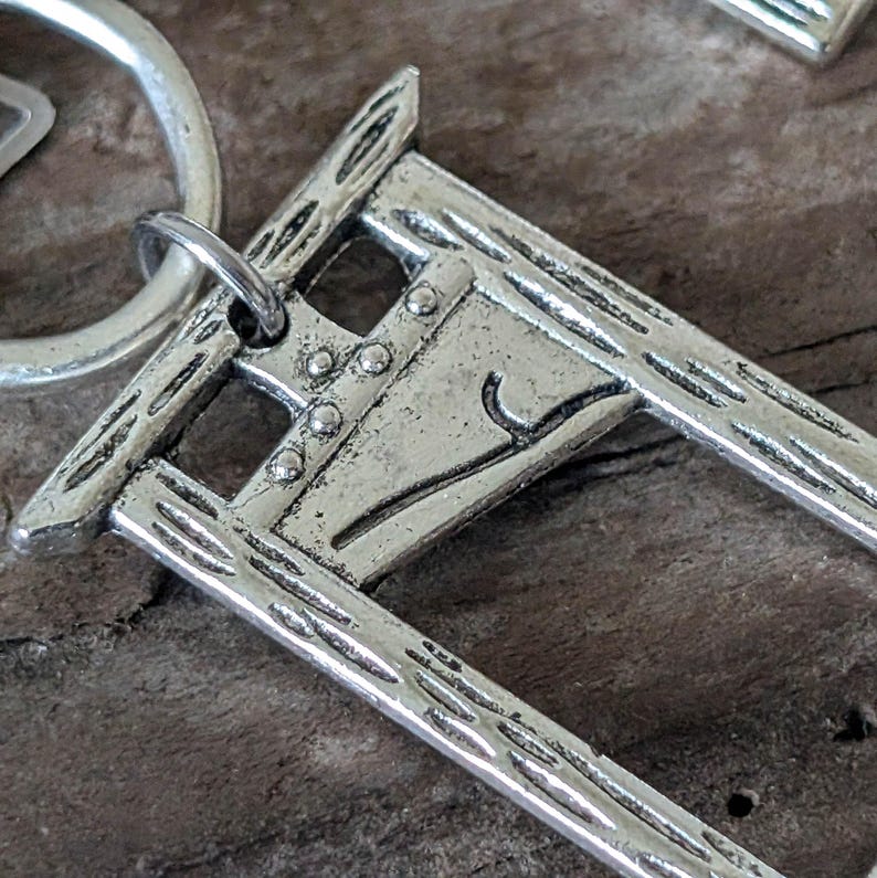 May include: A silver-colored keychain featuring a detailed miniature guillotine design. The keychain has a ring for attaching keys and a textured, aged appearance. The guillotine has a blade and a frame with rivets.