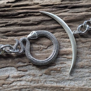 May include: A silver chain bracelet with a snake and crescent moon charm. The snake is coiled into a circle and the crescent moon is open at the top.