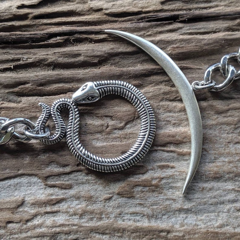 May include: A silver chain bracelet with a snake and crescent moon charm. The snake is coiled into a circle and the crescent moon is open at the top.