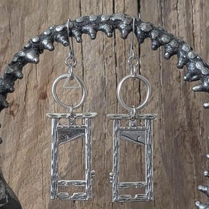 May include: Silver-toned earrings featuring a guillotine design. Each earring has a rectangular guillotine pendant with a detailed blade and frame, suspended from a circular and triangular accent. The earrings hang from a decorative, textured metal display.