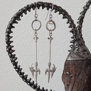 May include: A pair of silver earrings with a spearhead design. The earrings are hanging from a silver hoop and have a detailed spearhead design with a pointed tip.