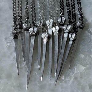 May include: A collection of necklaces featuring pendants shaped like metal nails. Each pendant has a textured, silver-toned head wrapped in thin wire, suspended from either a black or silver-toned metal chain. The nails have a pointed tip.